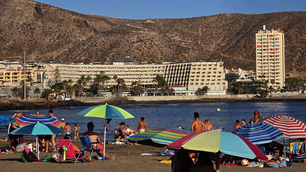 Słoneczna plaża w Los Cristianos na Teneryfie z plażowiczami, hotelami i górami w tle.
